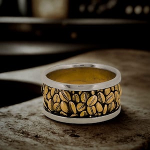 Op de afbeelding: Een zilveren ring met een gouden band, versierd met een patroon van koffiebonen. De ring heeft een gepolijste zilveren rand en een gouden binnenkant. Het koffiebonenontwerp geeft de ring een unieke uitstraling.