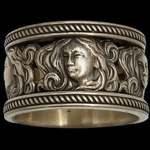 May include: A silver ring featuring a detailed design of three women's faces, each with flowing hair and intricate details. The ring has a rope-like border.