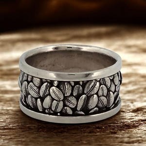 Op de afbeelding: Een zilveren ring met een band van gedetailleerde koffieboonontwerpen. De ring heeft een gepolijste afwerking en een brede band, met het koffieboonpatroon rond de omtrek. De ring is geplaatst tegen een houten achtergrond.
