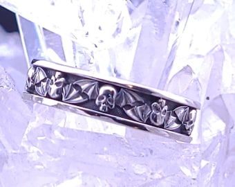 May include: A silver band ring with a skull and bat design. The ring is set against a white background.