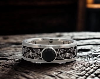 May include: A silver ring with a black gemstone center. The band features a repeating bat design in a dark gray color. The ring is set against a rustic wooden surface.