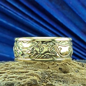 May include: A gold-colored ring with an intricate design featuring a running animal and swirling patterns. The ring is set against a blue background and rests on a wooden surface, showcasing its detailed craftsmanship.