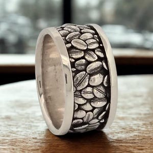 Op de afbeelding: Zilveren ring met een band van gedetailleerde koffieboonontwerpen. De ring heeft een gepolijste, gladde afwerking op de buitenste randen en het koffieboonpatroon is getextureerd.
