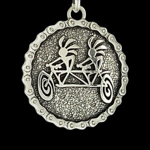 Puede incluir: Pendiente de plata con una imagen estilizada de dos personas montando una bicicleta tándem. El pendiente es circular y tiene un borde de eslabones de cadena.