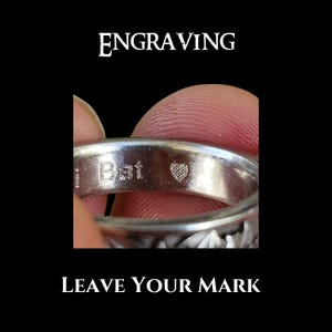 May include: Close-up of a silver ring with the word "Bat" engraved on it, along with a small heart symbol. The ring is held by a finger. The words "ENGRAVING" and "LEAVE YOUR MARK" are displayed in white text against a black background.