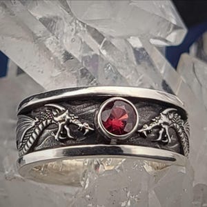 May include: A silver ring with a detailed dragon design on the band. The ring features a round, red gemstone in the center. The dragons are facing each other. The ring is set against a background of clear crystals.