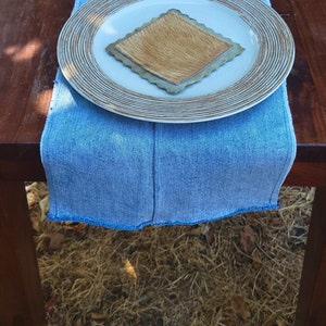 May include: A wooden table with a blue denim placemat and a white plate with a brown coaster on top. The table is set for a meal with a fork and a glass jar.