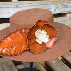 May include: A brown felt hat with a wide brim, decorated with a large brown leather flower and a smaller brown leather leaf. The hat is displayed on a black stand.