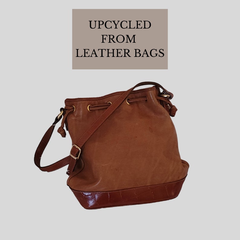 Op de afbeelding: Een bruine leren emmertassen met een trekkoord sluiting en een lange schouderband. De tas heeft een krokodil-gepr&auml;gd bodem en de tekst "UPCYCLED FROM LEATHER BAGS" is gedrukt op een bruin rechthoek.