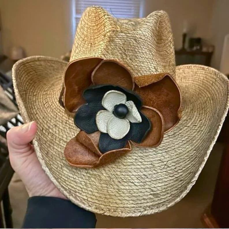 May include: A straw cowboy hat with a large brown leather flower accent. The flower has a white center with a black center and a black outer ring.