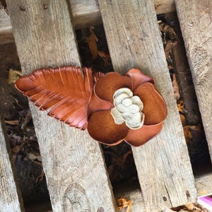Op de afbeelding: Een bruine leren bloem met een wit centrum en een bruin leren blad. De bloem en het blad liggen op een houten oppervlak.