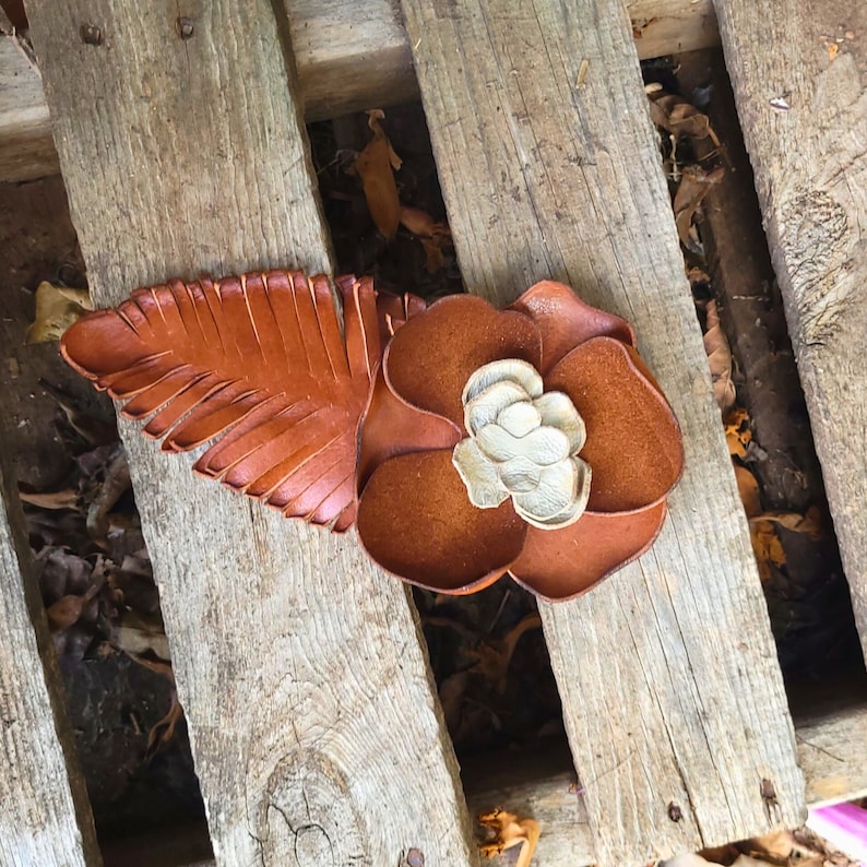 Op de afbeelding: Een bruine leren bloem met een wit centrum en een bruin leren blad. De bloem en het blad liggen op een houten oppervlak.