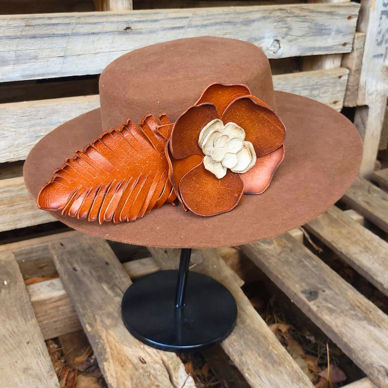 May include: A brown felt hat with a wide brim, decorated with a large brown leather flower and a smaller brown leather leaf. The hat is displayed on a black stand.