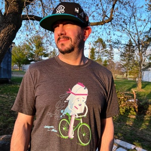 Puede incluir: Una persona lleva una gorra de béisbol negra y turquesa con un gráfico de un ojo y un pequeño gráfico en blanco y negro. La persona lleva una camiseta gris con un gráfico de dibujos animados de un helado en bicicleta.