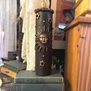 May include: A dark brown, cylindrical candle holder with a sun and star design. The candle holder is made of metal and has a gold-colored sun face and star accents. It is displayed on a stack of vintage books.