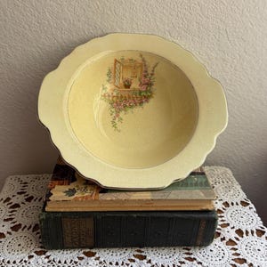 May include: A vintage yellow ceramic bowl with a scalloped edge and a gold rim. The bowl features a painted scene of a window box with flowers. The bowl is resting on top of two antique books.