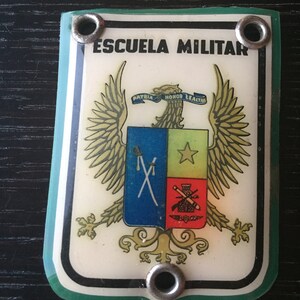 Op de afbeelding: Een wit en groen plastic badge met een gouden adelaar en schild. De badge heeft de tekst "ESCUELA MILITAR" en "PATRIA HONOR LEALTAD".