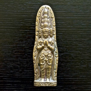 May include: A gold-coloured metal sculpture of a deity with multiple heads and hands, standing in a prayer position. The sculpture is detailed with intricate carvings and is set against a dark wooden background.