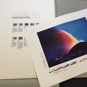 May include: A printed image of a colorful light show over a dark, rounded hill. The image is part of a larger publication titled "American Photographer".