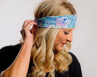 May include: A colorful headband with a floral and leaf pattern in shades of blue, pink, and white. The headband is worn by a person with blonde hair and a black shirt. The headband is a wide, stretchy fabric.