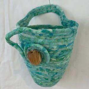May include: A teal blue fabric basket with a woven design and a wooden button. The basket has a handle and is hanging from a white wall.