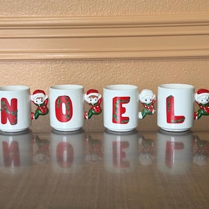 May include: Four white ceramic mugs spelling "NOEL" in red letters with a green leafy design. Each mug has a handle shaped like a Christmas elf. The mugs are on a reflective surface.