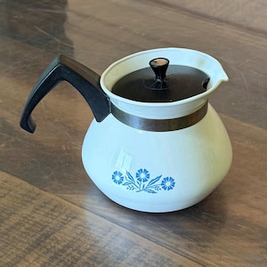 May include: A white teapot with a rounded body featuring a blue floral pattern. It has a black plastic handle and a matching black lid with a central knob. A metallic band encircles the upper portion of the teapot, connecting the handle to the body.