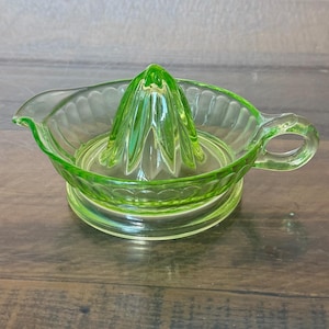 May include: A vibrant lime green glass citrus juicer featuring a central reamer cone, a circular basin with a scalloped rim, a small pouring spout on one side, and a sturdy handle on the opposite side, all crafted from translucent pressed glass.