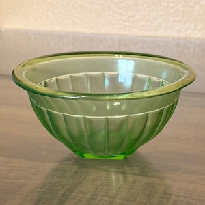May include: A translucent light green glass bowl featuring a rounded shape and a series of vertical fluted ridges along the exterior surface. The bowl has a smooth, thick rim and a clear, polished finish that allows light to pass through the glass.