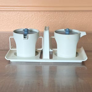 May include: A vintage cream-colored creamer and sugar bowl set with blue lids and handles. The set includes a creamer with a spout and a sugar bowl, both sitting on a matching tray. The set is made of plastic.