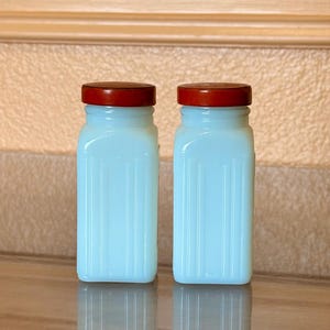 May include: Two matching light blue glass jars with ribbed sides and dark red lids. The jars are rectangular with rounded corners and sit on a reflective surface. The background is a light brown.