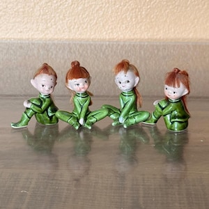 May include: Four ceramic figurines of elves with red hair and green outfits. The elves are sitting with their legs spread, and their hands are resting on their knees. The figurines have a glossy finish and are arranged in a row.