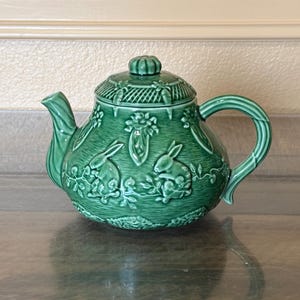May include: A green ceramic teapot with a lid, spout, and handle. The teapot features embossed floral and rabbit designs. The teapot is sitting on a reflective surface.