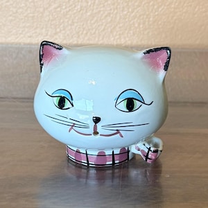 May include: A white ceramic cat figurine with black whiskers, blue eyes, and pink accents on the ears and collar. The cat has a whimsical design and a small handle on the side. The figurine is likely a decorative item.