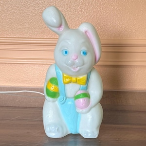May include: A white Easter bunny ornament with blue eyes, pink-lined ears, and a yellow bow tie. The bunny wears a light blue outfit and holds two decorated Easter eggs, one green and yellow, the other pink and green. A festive decoration.