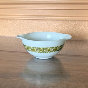 May include: A white Pyrex mixing bowl with a light green floral pattern around the rim. The bowl has two handles and is sitting on a wooden surface. The bowl is a light blue color.
