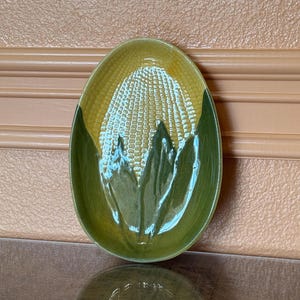 May include: A green and yellow ceramic serving dish shaped like an ear of corn. The dish has a raised design of corn kernels and husks.