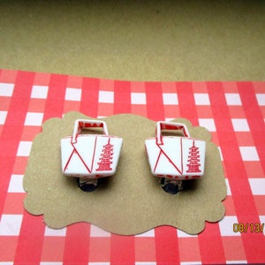 May include: Pair of white and red pagoda-themed clip-on earrings. Each earring features a miniature basket design with a red pagoda illustration and red accents. The earrings are displayed on a brown, scalloped card against a red and white checkered background.