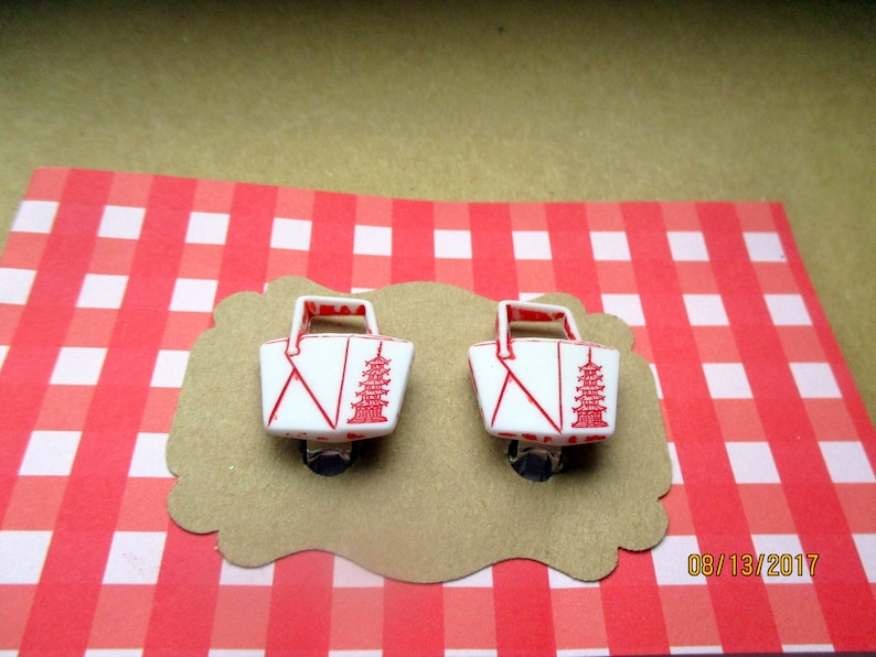May include: Pair of white and red pagoda-themed clip-on earrings. Each earring features a miniature basket design with a red pagoda illustration and red accents. The earrings are displayed on a brown, scalloped card against a red and white checkered background.