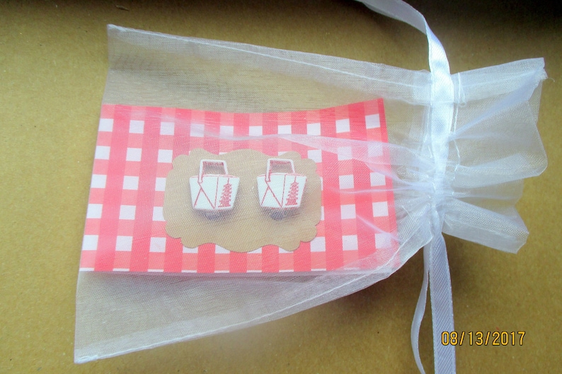 May include: A pair of white and red picnic basket earrings on a pink and white checkered background, presented in a sheer white gift bag. The earrings are on a brown, scalloped-edge card. The bag is tied with a white ribbon.