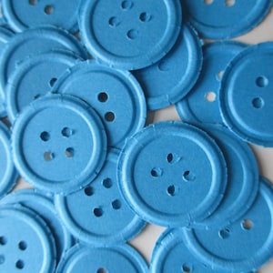Puede incluir: Un conjunto de botones redondos de color azul claro, cada uno con cuatro agujeros. Los botones tienen una superficie ligeramente texturizada y un borde exterior en relieve. Estos suministros de manualidades son adecuados para coser, hacer álbumes de recortes u otros proyectos.
