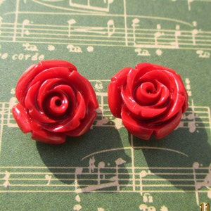 May include: Two bright red rose-shaped cabochons. The roses are highly detailed with multiple layers of petals. The cabochons are set against a green background with musical notes.