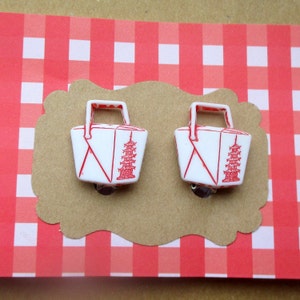 May include: A pair of white and red earrings shaped like takeout containers. Each earring features a red pagoda design and a red handle. The earrings are displayed on a brown, scalloped card against a red and white checkered background.