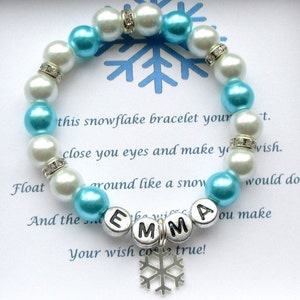 Könnte beinhalten: Ein Armband mit weißen und hellblauen Perlen, mit silbernen Strass-Abstandshaltern. Das Armband hat den Namen "EMMA" in silbernen Buchstabenperlen und einen Schneeflocken-Anhänger. Das Armband wird auf einer Karte mit Text präsentiert.