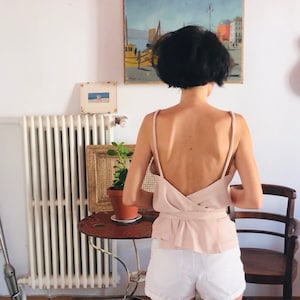 以下が含まれることがあります： 女性は、背中にラップディテールのある薄いピンクのノースリーブトップを着ています。トップはウエストで結ばれています。彼女は白いデニムのショートパンツも履いています。
