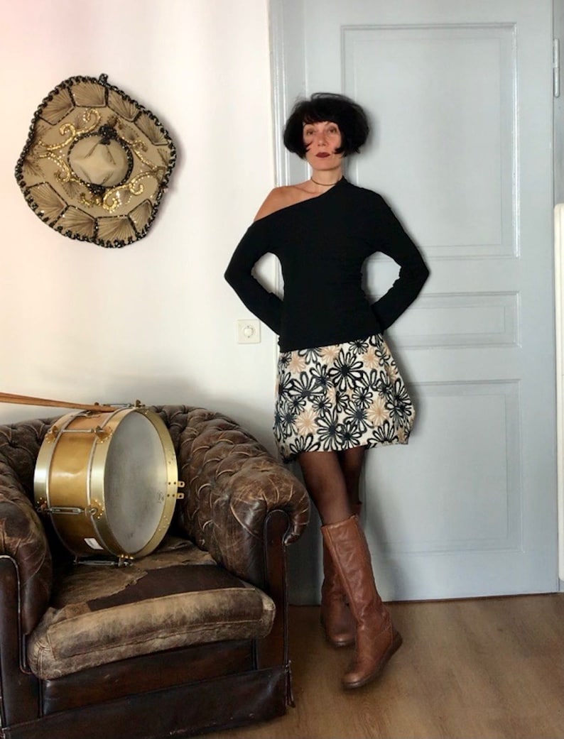 Puede incluir: Una mujer con una camisa negra de manga larga con un dise&ntilde;o de un solo hombro, una falda de estampado floral y botas marrones de ca&ntilde;a alta. Est&aacute; de pie en una habitaci&oacute;n con un tambor vintage y un gran sombrero en la pared.