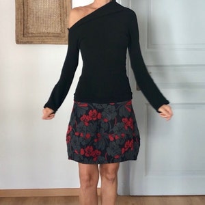 Puede incluir: Una camisa negra de manga larga con un escote de un hombro y una falda de estampado floral. La falda es negra con flores rojas.