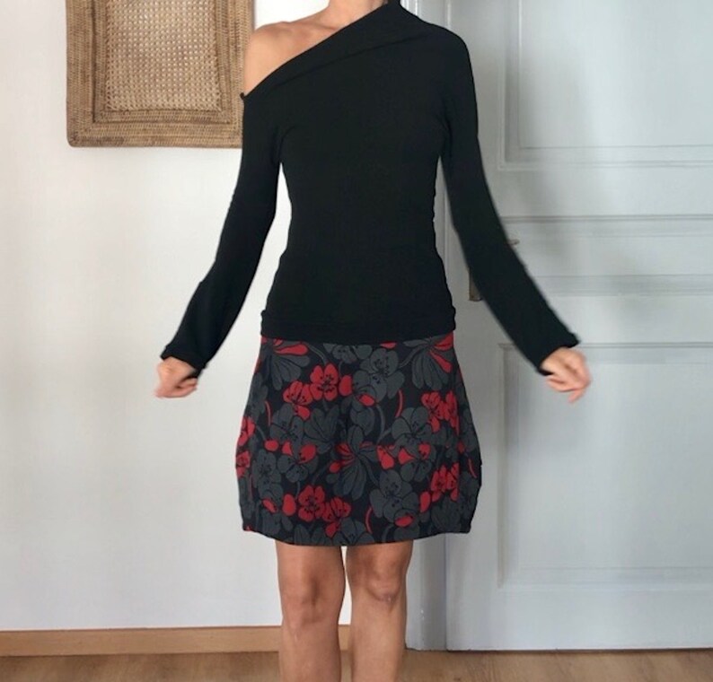 Puede incluir: Una camisa negra de manga larga con un escote de un hombro y una falda de estampado floral. La falda es negra con flores rojas.