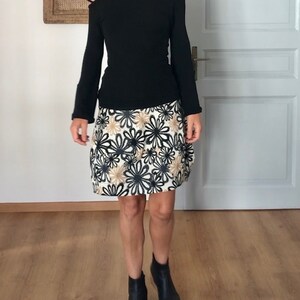 Puede incluir: Una camisa negra de manga larga con un escote de un solo hombro combinada con una falda blanca con estampado floral negro y dorado. La falda es de largo hasta la rodilla y tiene una silueta ligeramente acampanada. La persona lleva botines negros de tobillo.