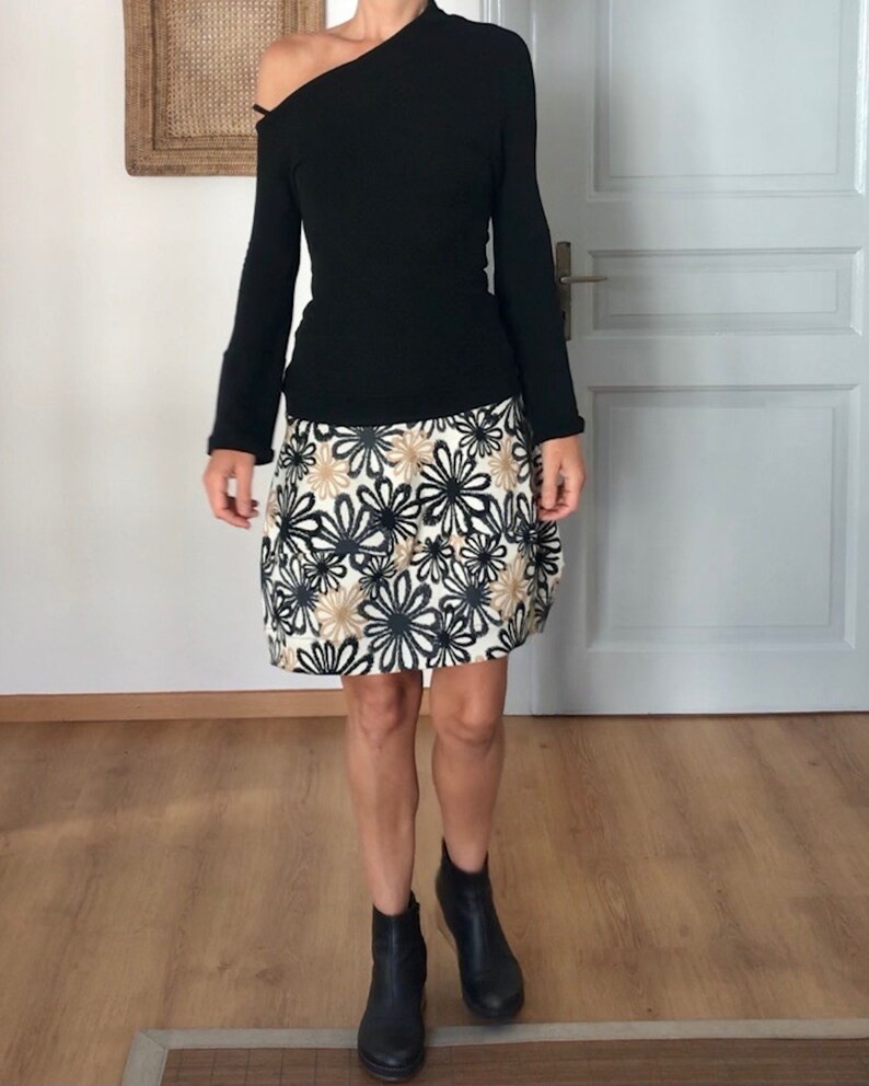 Puede incluir: Una camisa negra de manga larga con un escote de un solo hombro combinada con una falda blanca con estampado floral negro y dorado. La falda es de largo hasta la rodilla y tiene una silueta ligeramente acampanada. La persona lleva botines negros de tobillo.